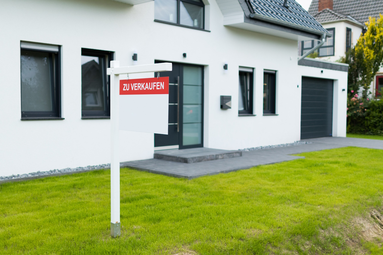 Schild mit der Aufschrift “Zu verkaufen” steht vor einem weißen Einfamilienhaus in Schleswig-Holstein, da der Eigentümer die Immobilie verkaufen möchte.